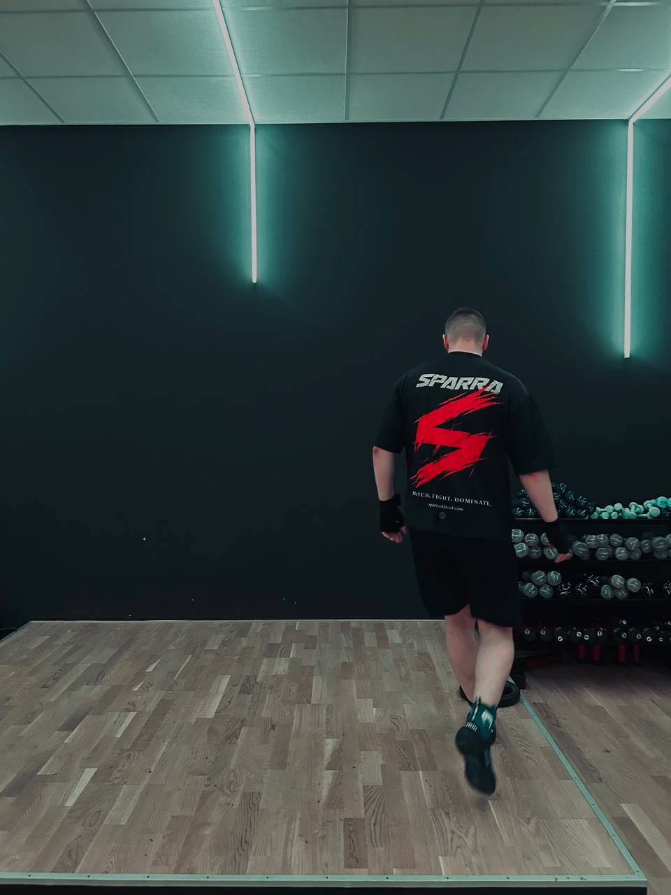 Boxer walking through a boxing gym wearing the Sparra Fight Tee — back view with heavy bag and ring ropes in the background
