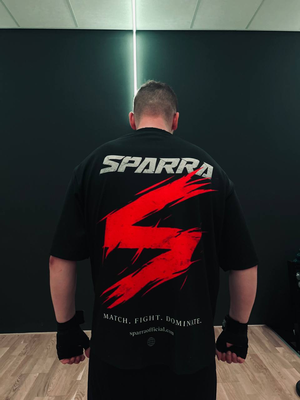 Boxer in a gym wearing the Sparra Fight Tee — back view with SPARRA slash graphic and 'Match. Fight. Dominate.' tagline